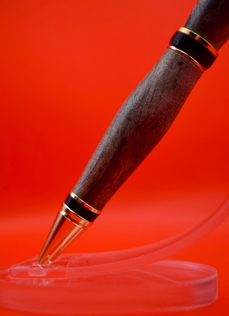 May include: A handcrafted wooden pen with a dark brown finish, accented by gold-colored metal bands and a gold-colored tip. The pen is displayed against a vibrant red background.