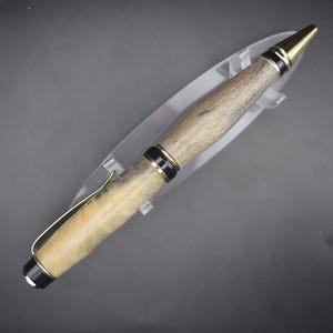 May include: A wooden pen with gold and black accents. The pen has a light brown and beige wood grain barrel, a gold-colored clip, and a gold-colored tip. The pen is displayed on a clear plastic stand against a dark background.