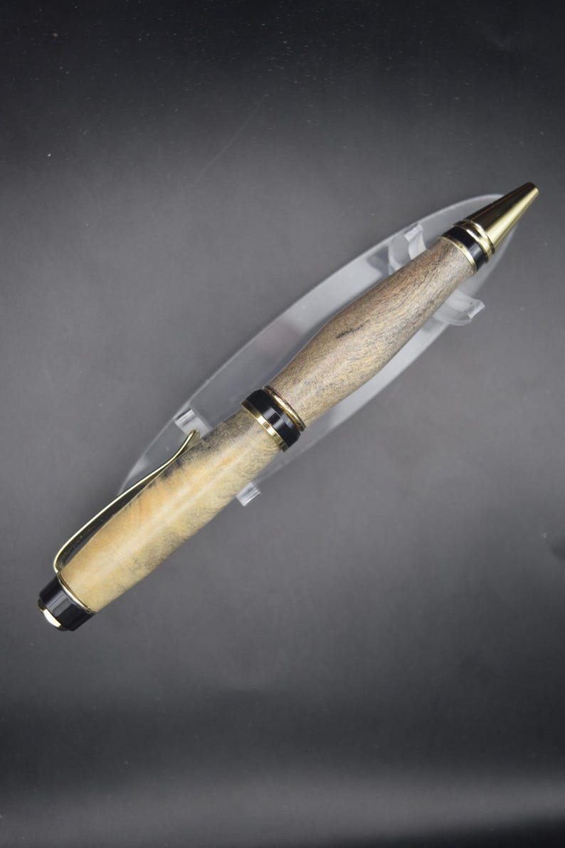 May include: A wooden pen with gold and black accents. The pen has a light brown and beige wood grain barrel, a gold-colored clip, and a gold-colored tip. The pen is displayed on a clear plastic stand against a dark background.