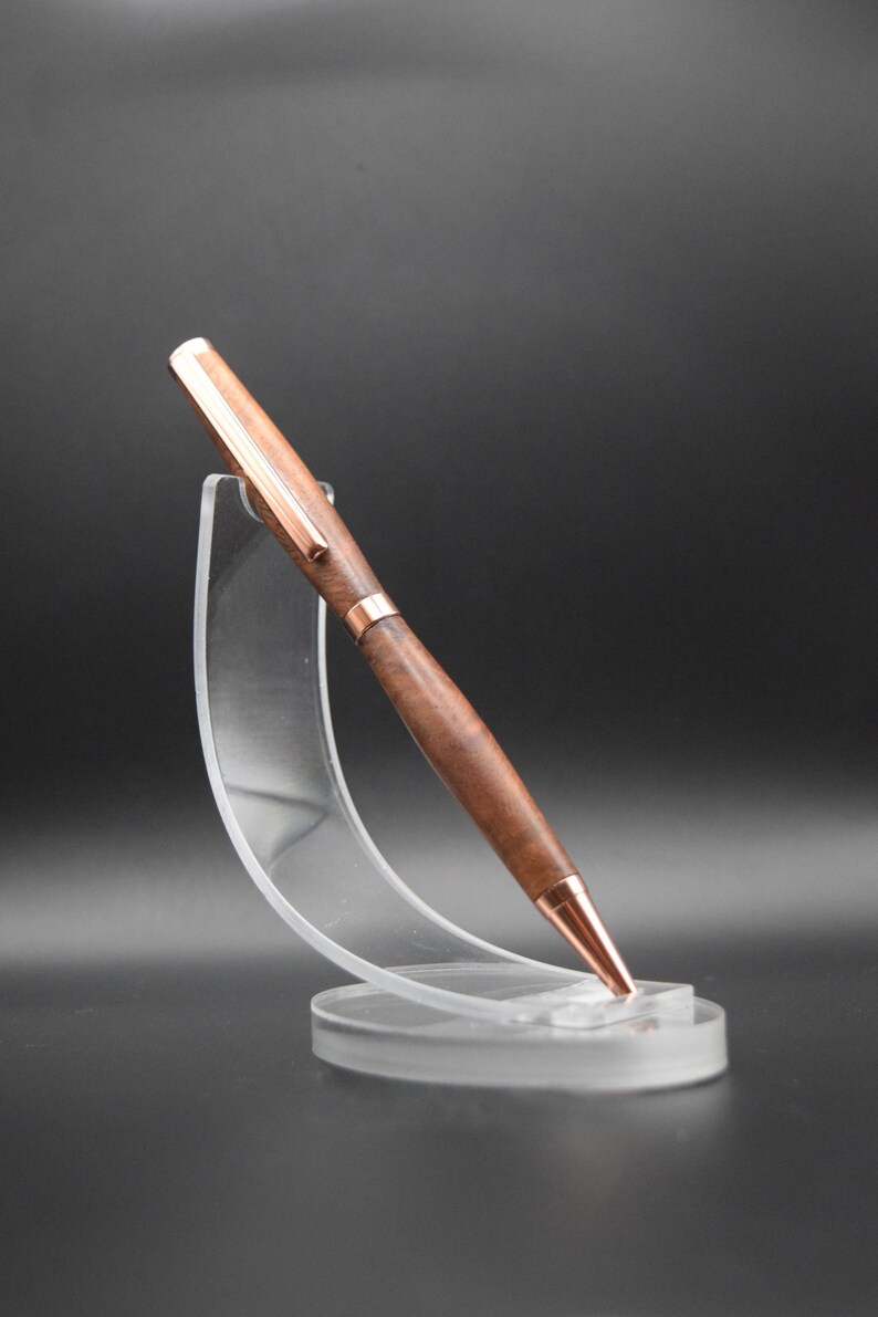 May include: A wooden pen with a copper colored top and bottom. The pen is resting on a clear plastic stand.