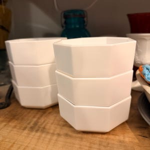 May include: Two stacks of three white hexagonal bowls made of a smooth, solid material. Each bowl features six distinct flat sides and a clean, uniform finish. The bowls are arranged in two vertical columns, showcasing their geometric shape.