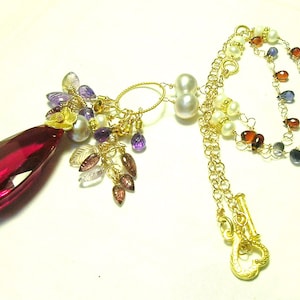 May include: A gold necklace with a large, teardrop-shaped, red gemstone pendant. The pendant is surrounded by smaller gemstones in shades of purple, pink, and white. The necklace also features a gold chain with a heart-shaped clasp.
