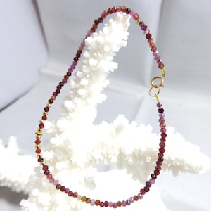 May include: A delicate necklace featuring small, faceted beads in shades of red, pink, and purple. The beads are strung together with gold-colored spacers and a gold clasp. The necklace is displayed against a white background.