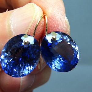 May include: A pair of oval-shaped, faceted blue gemstones suspended from gold-toned metal ear wires. The stones exhibit a deep, translucent blue color with intricate light-reflecting facets across their surfaces, creating a brilliant appearance.