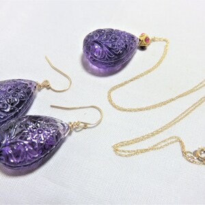 May include: A set of amethyst earrings and a necklace. The earrings are teardrop shaped and feature intricate carvings. The necklace has a teardrop shaped pendant with carvings and a small pink stone accent. The chain is gold.