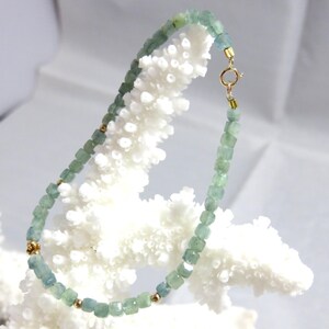 May include: A delicate necklace featuring small, square-cut aquamarine beads in shades of blue-green. The necklace is accented with gold-colored beads and a clasp. The jewelry is displayed on a white, textured surface, creating a contrast with the cool tones of the gemstones.