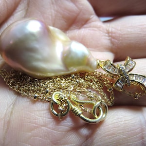 Puede incluir: Un gran colgante de perla irregular, iridiscente, de color blanco roto con una cadena de oro y un dije de lazo de oro con cristales transparentes.