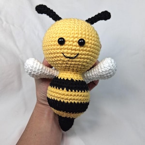 Puede incluir: Juguete de ganchillo hecho a mano con forma de abeja. Tiene un cuerpo amarillo con rayas negras y alas blancas. La abeja tiene ojos negros, antenas y una cara sonriente. El juguete mide aproximadamente 13 cm de alto.