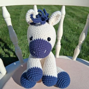 May include: A handmade crochet stuffed animal in the shape of a horse. The toy is primarily off-white with navy blue accents on the muzzle, hooves, and mane. The horse has black button eyes and is sitting on a pink cushion.