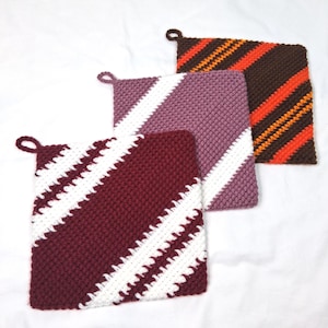 Può includere: Tre presine quadrate fatte a mano, nei toni del bordeaux, lavanda e marrone. Ogni presina presenta un motivo a righe diagonali in colori contrastanti. La presina bordeaux ha strisce bianche, la lavanda ha bianche e la marrone ha arancioni.