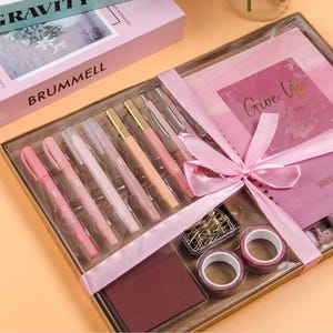 May include: A gift set with pink and gold pens, a notebook, washi tape, and paperclips. The set is tied with a pink ribbon and includes the text "Give Up". The box is labeled "BRUMMELL" and "GRAVITY".