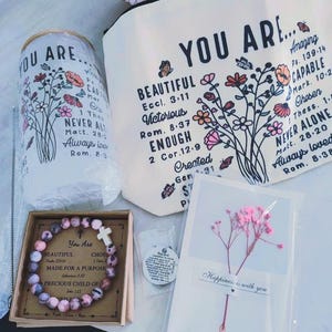 May include: A gift set with a pink and white bracelet with a cross charm, a white canvas bag with floral designs and the text "You Are... Beautiful, Victorious, Enough, Created", a white card with pink dried flowers and the text "Happiness is with you", and a small white round charm with a cross.