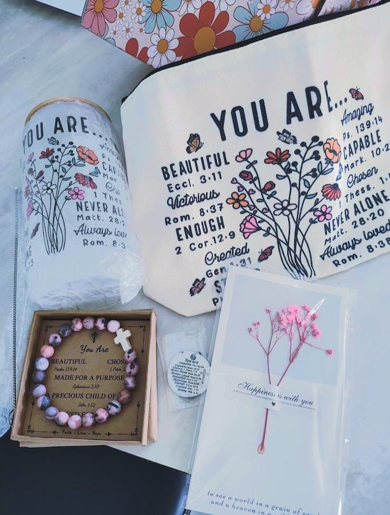 May include: A gift set with a pink and white bracelet with a cross charm, a white canvas bag with floral designs and the text "You Are... Beautiful, Victorious, Enough, Created", a white card with pink dried flowers and the text "Happiness is with you", and a small white round charm with a cross.