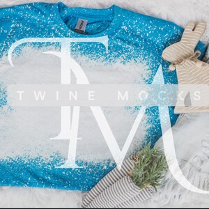 May include: A blue t-shirt with white bleach splatters. The shirt is laying on a white fuzzy surface with a pair of light blue denim shorts and a wooden bunny figurine.