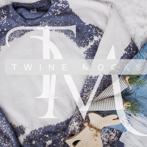 Puede incluir: Una sudadera blanca con un diseño de salpicaduras de lejía azul. La sudadera está sobre una superficie blanca y peluda con un par de pantalones vaqueros azules y un adorno de madera.