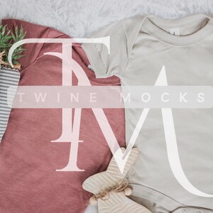 May include: Two baby onesies, one in a dusty rose colour and one in a light beige colour, laid out on a white fluffy surface. The onesies are blank and ready for customisation. The text "TWIN MOCKS" is printed on the onesies.