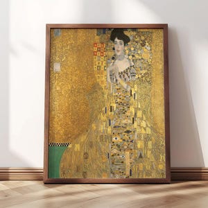 May include: Framed print of a Gustav Klimt painting. The artwork features a woman in a gold dress with intricate patterns and details. The frame is a dark brown color, and the background is a golden hue. The artwork is displayed on a white wall.