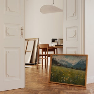 May include: A framed landscape painting of a green meadow with yellow wildflowers and a mountain range in the background. The painting is hanging on a wall in a room with wooden floors.