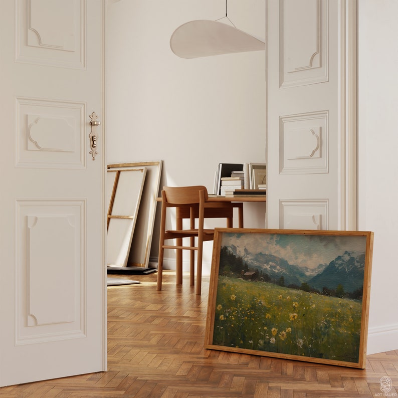 May include: A framed landscape painting of a green meadow with yellow wildflowers and a mountain range in the background. The painting is hanging on a wall in a room with wooden floors.