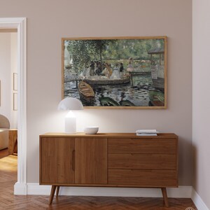 Op de afbeelding: Een houten dressoir met twee deuren en drie lades. Het dressoir is lichtbruin van kleur en heeft een modern ontwerp. Een witte lamp en een witte kom staan op het dressoir. Een ingelijst schilderij van een groep mensen op een meer hangt aan de muur boven het dressoir.