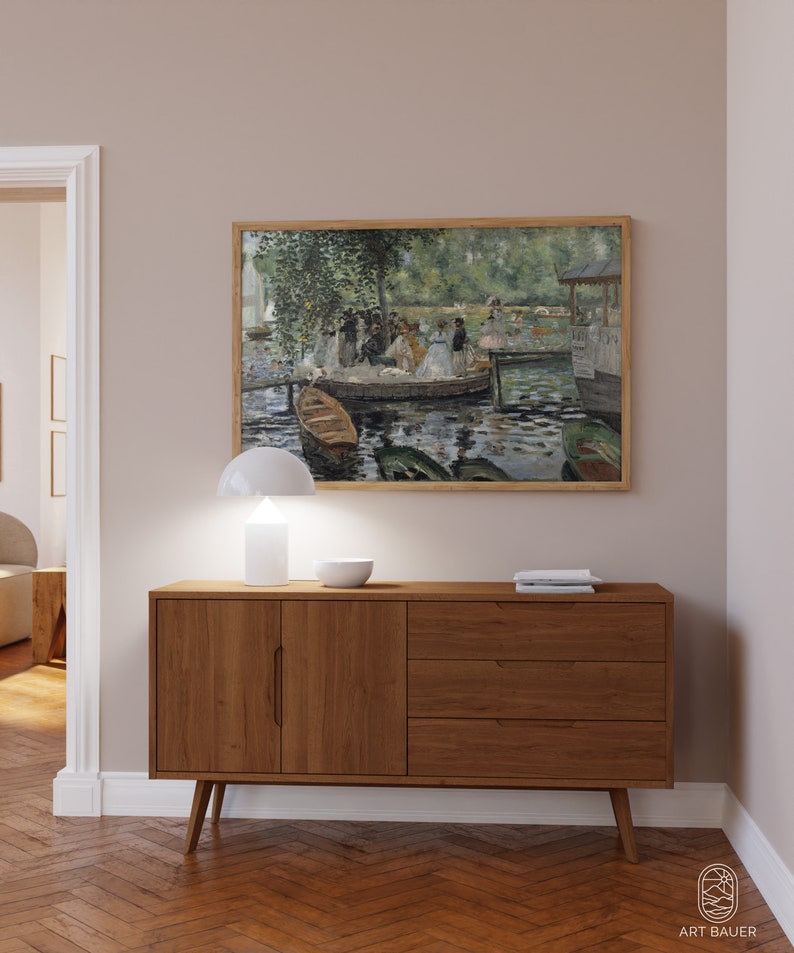 Op de afbeelding: Een houten dressoir met twee deuren en drie lades. Het dressoir is lichtbruin van kleur en heeft een modern ontwerp. Een witte lamp en een witte kom staan op het dressoir. Een ingelijst schilderij van een groep mensen op een meer hangt aan de muur boven het dressoir.