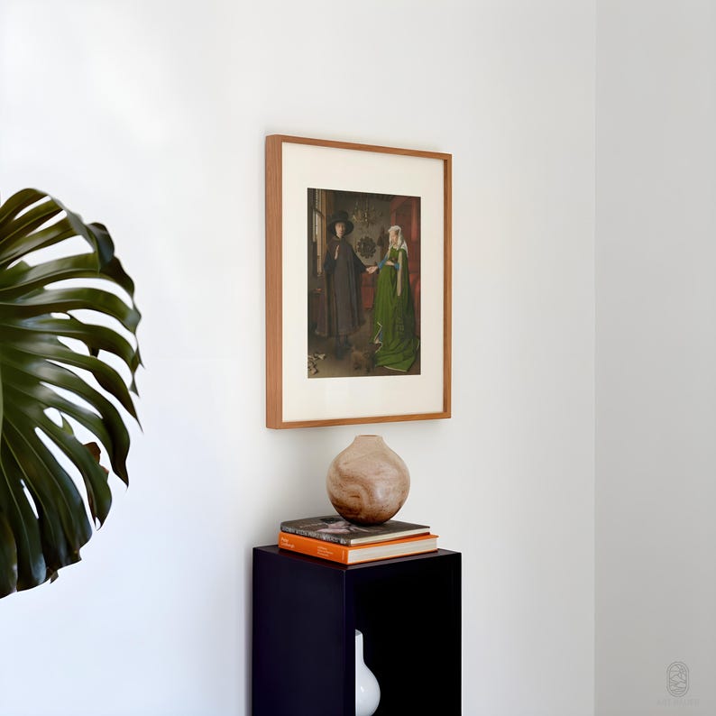 Puede incluir: Una obra de arte enmarcada que presenta una pintura hist&oacute;rica de dos figuras con vestimenta de &eacute;poca. La obra de arte est&aacute; en un marco de madera marr&oacute;n claro y cuelga de una pared blanca. Debajo, un mueble azul oscuro con un jarr&oacute;n decorativo y libros.