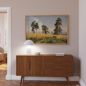 May include: A wooden sideboard with two doors and three drawers. The sideboard is a light brown color and has a modern design. A white lamp and a white bowl are on top of the sideboard. A framed landscape painting of a field with trees is hanging on the wall above the sideboard.