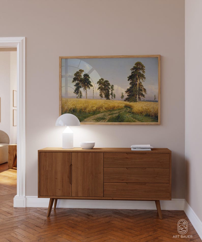 May include: A wooden sideboard with two doors and three drawers. The sideboard is a light brown color and has a modern design. A white lamp and a white bowl are on top of the sideboard. A framed landscape painting of a field with trees is hanging on the wall above the sideboard.