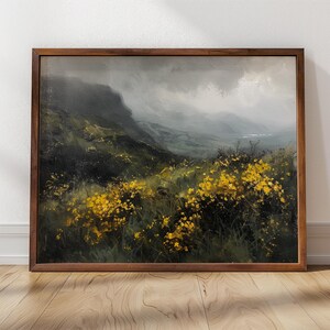 Puede incluir: Una pintura enmarcada de un paisaje brumoso con una ladera cubierta de flores silvestres amarillas. La pintura está colocada contra una pared blanca con un suelo de madera.