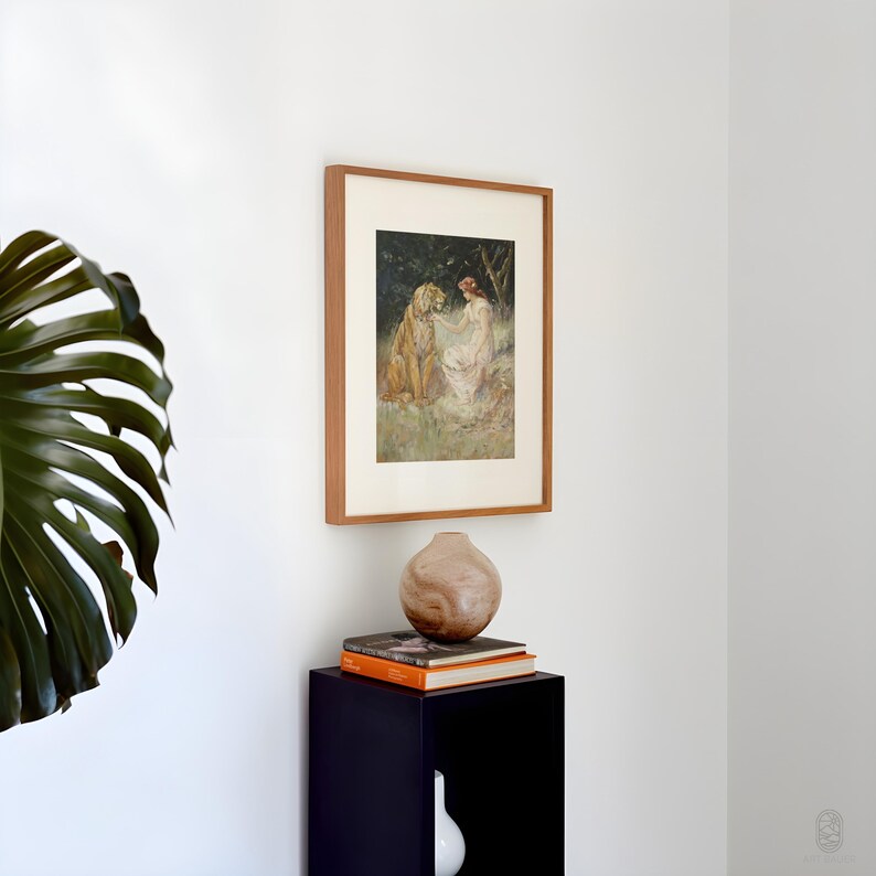 May include: A framed painting of a woman in a white dress sitting with a tiger in a forest setting. The painting is on a black shelf with a wooden vase and books.