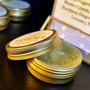 May include: A gold-colored tin of herbal vapor rub with the lid slightly ajar, revealing a light green balm. The label reads "HERBAL VAPOR RUB". Other similar tins are visible in the background. The product is likely for aromatherapy or topical use.