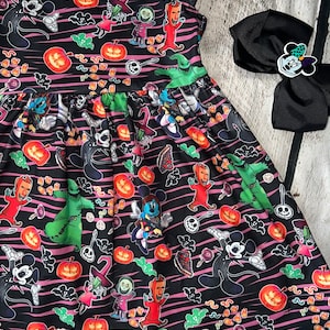 Può includere: Abito nero con stampa a tema Halloween con personaggi, zucche e caramelle. L'abito ha uno scollo tondo e maniche corte. È visibile anche un fiocco nero con un design di Minnie Mouse.