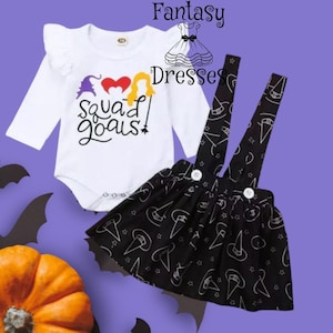 Könnte beinhalten: Ein zweiteiliges Baby-Outfit für Halloween. Es besteht aus einem weißen Langarm-Body mit dem Text "Squad Goals!" in Schwarz, Lila, Rot und Gelb. Dazu gehört ein schwarzer Rock mit weißem Hexenhut- und Sternenmuster.
