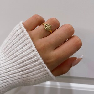 May include: A gold ring with a textured circular design. The ring is on a finger with a light pink nail polish.