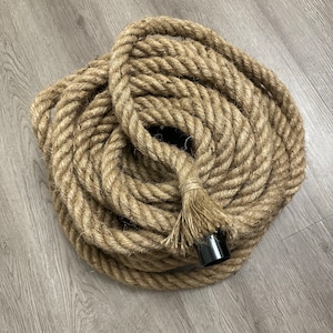 May include: Coiled natural fibre rope with a black plastic end piece. The rope is thick and appears to be made of jute or a similar material. The rope is lying on a light grey wooden surface.