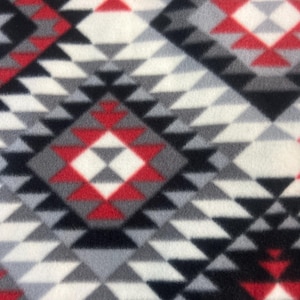 May include: Close-up of a fleece fabric with a geometric pattern. The design features repeating diamond shapes in red, black, white, and gray. The fabric has a soft, textured appearance, suitable for blankets or apparel.