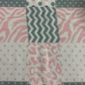 Puede incluir: Primer plano de una tela suave estampada. El diseño presenta un mosaico de cuadrados blancos, rosas y grises. Algunos cuadrados tienen rayas de cebra rosas, otros zigzags grises y algunos manchas de leopardo rosas.