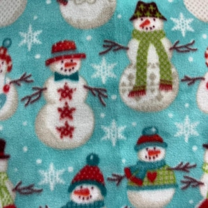May include: A repeating pattern of snowmen on a light blue background. The snowmen have various hats, scarves, and bow ties in red, green, and blue. White snowflakes are scattered throughout the design. The snowmen have twig arms and carrot noses.