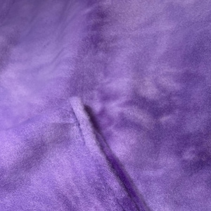 May include: Close-up of a soft, plush fabric in shades of purple. The material has a velvety texture with subtle variations in color, creating a cloud-like appearance. The fabric is folded, revealing a seam.