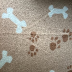 May include: Tan fleece fabric featuring a repeating pattern of white dog bones and brown paw prints. The soft material is suitable for pet bedding or craft projects.