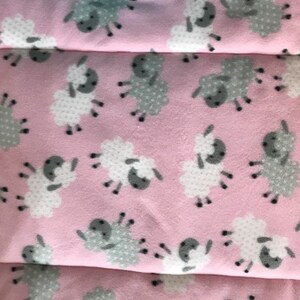 May include: Pink fabric featuring a pattern of white and grey sheep. The sheep have white bodies with grey faces and are scattered across the fabric. The fabric has a quilted border.