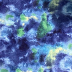 May include: A close-up of a soft, textured fabric with a vibrant, abstract pattern. The design features a blend of deep blues, light blues, and touches of green and yellow, creating a cloudy or watercolor effect. The fabric appears to be fleece or a similar material.