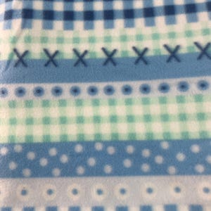 May include: A close-up of a blue and white patterned fabric. The design includes stripes with various patterns: polka dots, cross-stitches, and checkered squares in shades of blue and white. The fabric appears soft and textured.