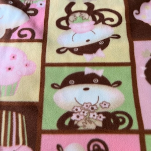 May include: A close-up of a patterned fabric featuring a repeating design of cartoon monkeys in a grid. The monkeys are brown, white, and pink, set against a backdrop of pink, green, and yellow squares, with brown borders.