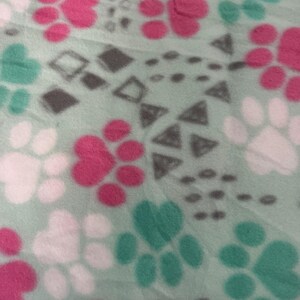 May include: A light green fleece fabric with a repeating pattern of pink, white, and teal paw prints. The design also includes gray geometric shapes, such as diamonds and triangles, scattered across the surface. The fabric has a soft, plush texture.