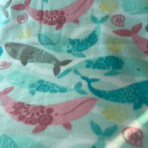 May include: A light blue fabric with a pattern of whales in shades of pink, teal, and gray. The whales are swimming in different directions, with some featuring textured details. Scattered yellow and pink sea life elements complete the design.