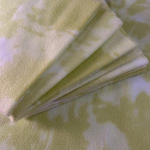 May include: A pile of soft, tie-dye fleece blankets in pale yellow and white. The blankets are folded, showing the fabric's texture and pattern. The overall look is cosy and inviting, perfect for snuggling up with.