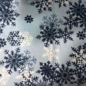 May include: A light blue fleece fabric with a repeating pattern of snowflakes in shades of navy and white. The snowflakes vary in size and are evenly distributed across the material, creating a winter theme.