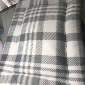 May include: A grey and white plaid dog bed with a white border and a small blue paw print label that says "Sherrie's Comforts".