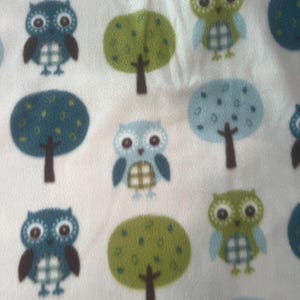 May include: White fabric with a repeating pattern of cartoon owls and trees. The owls are in shades of blue, green, and brown, with large eyes and patterned bellies. The trees are green and blue with brown trunks.